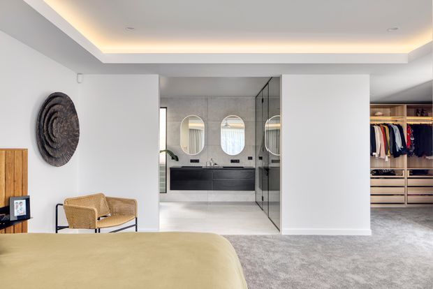 A bright contemporary bedroom with a pale upholstered bed in the foreground, opening directly onto a refined bathroom and a walk-in wardrobe. The bathroom features a dark floating vanity, twin rounded mirrors, pale stone or tiled surfaces, and a frameless glass shower enclosure. A recessed ceiling with concealed warm lighting, soft grey carpet, white walls, timber accents, and woven seating create a calm, polished interior.