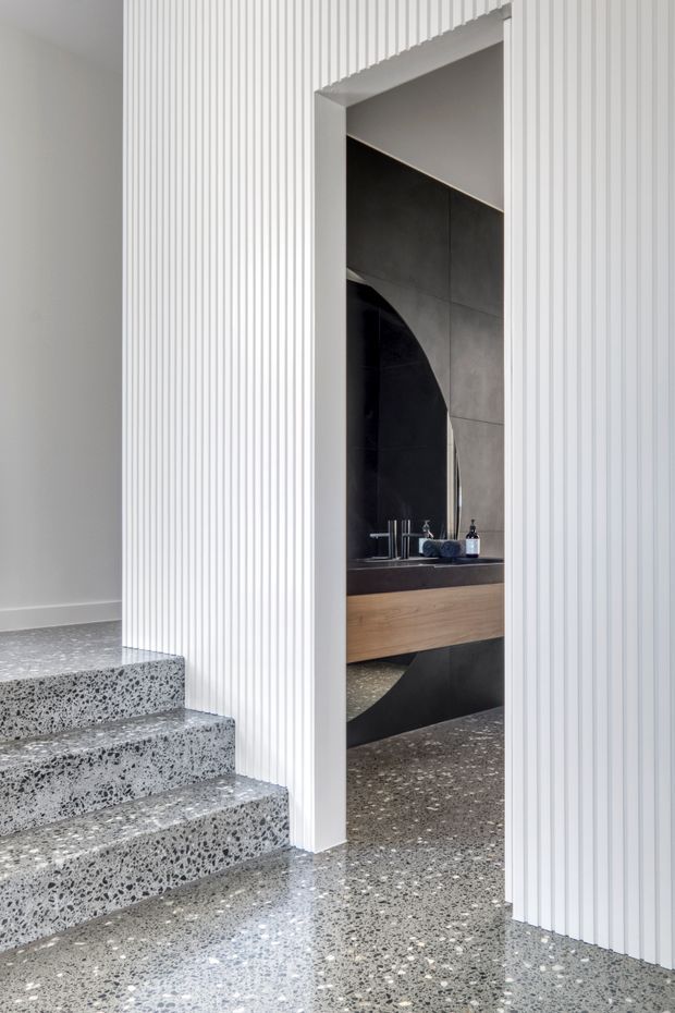 Contemporary interior view showing a terrazzo-finished stairway and floor beside a bathroom or powder room. The space features white vertically fluted wall panelling, a dark tiled feature wall with a large arched mirror, and a floating timber vanity with a dark benchtop and basin fixtures.