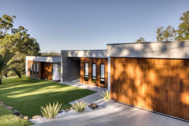 Single-storey contemporary residence with a low, flat-roofed composition, broad concrete-rendered walls, warm vertical timber garage cladding, and narrow dark-framed windows. The house is set within a landscaped suburban garden with lawn, agave plantings, gravel edging, and a concrete driveway, photographed in clear daylight.