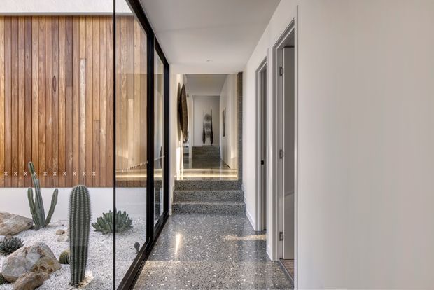 A bright contemporary residential hallway with polished terrazzo flooring, white walls, and several grey-trimmed doorways. Full-height black-framed glazing runs along the left side, looking onto an indoor cactus courtyard with a vertical timber feature wall. Steps lead toward a further corridor with artwork and a console visible in the distance.