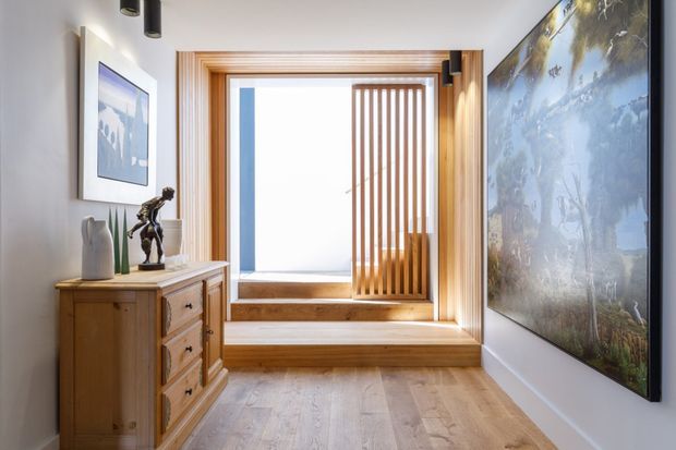 Bright, refined residential hallway or entry space featuring a large timber-framed window seat with vertical timber screening, pale timber flooring, a traditional timber chest of drawers, framed artwork, and a large nature-themed wall mural. The space combines warm natural wood with white walls and abundant daylight.