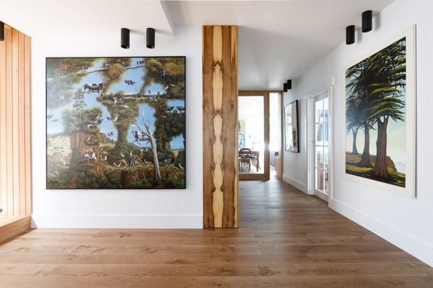 A bright contemporary hallway or gallery-like circulation space with wide timber flooring, white walls, and extensive framed artwork. A prominent natural timber structural or decorative post divides the corridor, while black cylindrical ceiling spotlights provide focused illumination. Glazed doors and windows line the right side, with a dining area visible at the far end. The space has a warm, curated residential character.