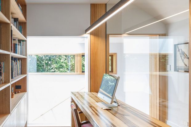 A bright, contemporary home office with a long timber desk, desktop computer, built-in open shelving filled with books, and integrated timber cabinetry. Full-height glazed partitions and interior windows bring in abundant natural light, while linear recessed and suspended lighting emphasizes the clean geometry. The palette is predominantly white with warm natural timber.