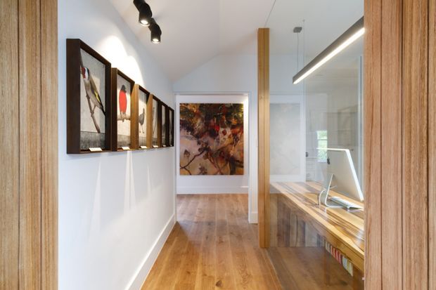 A narrow, bright timber-lined hallway or home office passage with warm timber flooring, vertical timber framing, framed artwork along the wall, and a built-in timber desk visible behind a glass partition. Contemporary linear lighting and clean white walls create a refined, gallery-like workspace connection.