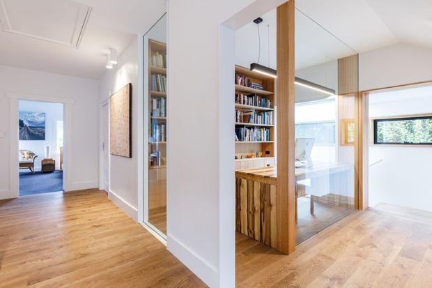 Bright contemporary residential hallway with warm timber flooring, white walls and ceiling, and integrated timber-and-glass joinery. A built-in bookshelf and timber cabinet frame a glazed opening into a light-filled living or study area, while a doorway at the far end opens to a room with mountain views. Clean-lined detailing, recessed lighting, and full-height glazing create an airy, refined interior.