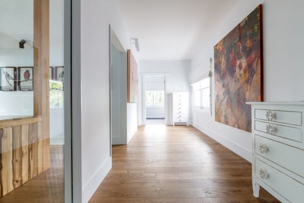 Bright, elongated residential hallway or landing with white walls, warm timber flooring, multiple internal doorways, and abundant natural light from windows at the far end and along the right wall. A large colourful artwork and a pale traditional chest of drawers provide visual accents. A glazed opening on the left reveals timber cabinetry and shelving in an adjacent room.