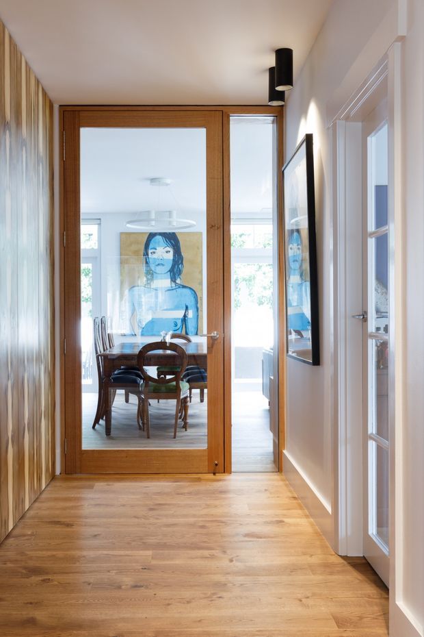 A bright residential hallway with warm timber flooring and a full-height timber-framed glazed door leading into a dining room. The dining area contains a round wooden table and chairs, with a large blue-toned artwork visible beyond and reflected in a wall-mounted mirror. White walls, white-trimmed internal doors, timber wall detailing, and black cylindrical ceiling lights create a refined, contemporary interior.