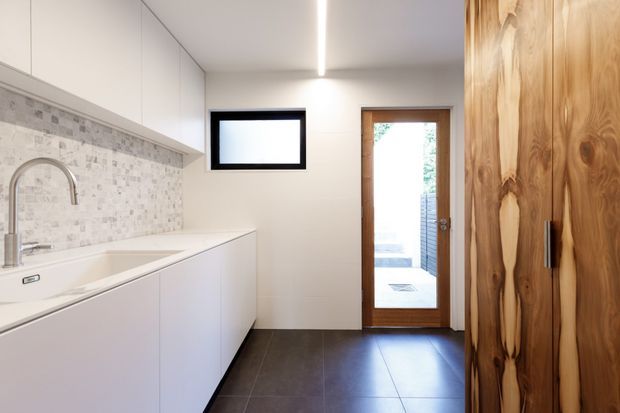 A bright, contemporary galley-style laundry with streamlined white full-height cabinetry, a long white benchtop and integrated sink, pale mosaic tile splashback, dark large-format tiled flooring, and a warm natural timber door and tall storage doors. A narrow linear ceiling light and small high-level window provide additional illumination. A glazed door opens to an outdoor area visible beyond.