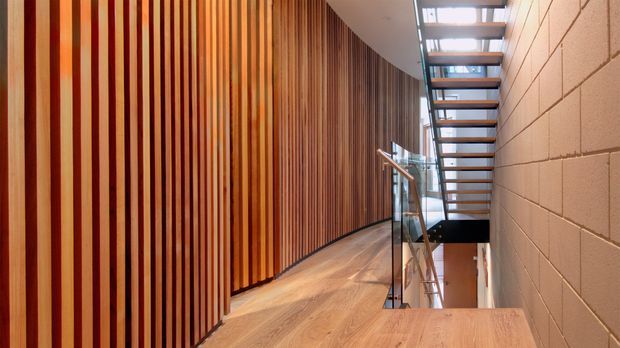 Interior view of a contemporary stairway and landing. A curved wall is clad in vertical warm timber battens, contrasting with a pale exposed block wall. The timber floor continues to a stair landing beside a glass balustrade, with an open-riser timber-and-metal staircase and skylight above. The space is bright, warm, and architecturally detailed.