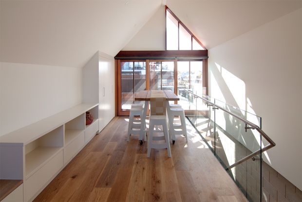 A bright contemporary mezzanine-level dining area with a long timber dining table and white stools, set beside full-height glazing and a triangular clerestory window. Warm timber flooring, white built-in cabinetry, exposed dark timber window framing, and a glass balustrade overlooking the lower level create an airy double-height interior.