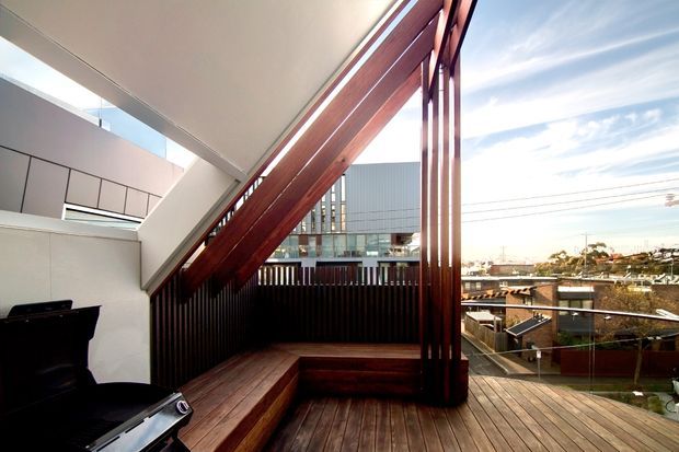 A contemporary elevated timber deck or terrace adjoining a modern building, framed by strong reddish-brown timber beams and vertical screening. Built-in timber bench seating lines the edge, with a compact outdoor grill or barbecue at the left. The open view overlooks a suburban urban setting, while the crisp geometry, glazing, and exposed structural framing create a striking architectural composition.