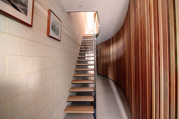 A contemporary interior stairway with open-riser timber treads and slim dark metal supports, rising between a pale exposed concrete block wall and a dramatic curved wall of vertical timber battens. The narrow circulation space has a polished grey floor, recessed ceiling lights, framed artwork, and bright glazing at the upper landing.