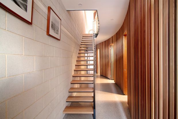 Interior view of a narrow contemporary stairway and hallway. Open-riser timber stairs with a slim metal handrail rise beside a painted masonry block wall, while the curving corridor is lined with vertical timber battens and integrated doors. Framed artwork and bright natural light add detail to the warm, refined circulation space.