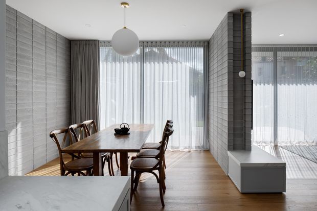 A bright, minimalist dining area with a rectangular timber dining table and six wooden chairs, positioned beside floor-to-ceiling sheer curtains. Grey block-patterned wall surfaces frame the room, with a sculptural spherical pendant light overhead and a low white built-in cabinet beneath a dark vertical column. Part of a pale stone kitchen island is visible in the foreground. The space has a restrained contemporary aesthetic with natural timber flooring and abundant filtered daylight.