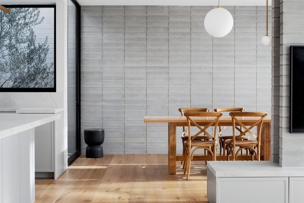 Bright contemporary dining area viewed from an adjoining kitchen or living space. The room features a timber dining table with matching cross-back timber chairs, warm timber flooring, a pale grey horizontally lined masonry or panelled feature wall, white built-in cabinetry, black-framed glazing, and spherical pendant lights. A partial television and white media cabinetry are visible at right, while a large window brings in views of foliage.