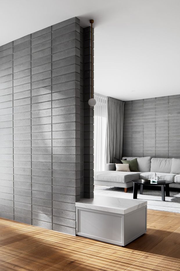 A bright contemporary living room featuring exposed grey concrete block walls, a light upholstered sectional sofa, black coffee table, floor-to-ceiling curtains, and warm timber flooring. A partial-height concrete block partition creates separation within the open-plan space, with a minimal globe pendant and strong sunlight casting linear shadows across the floor.