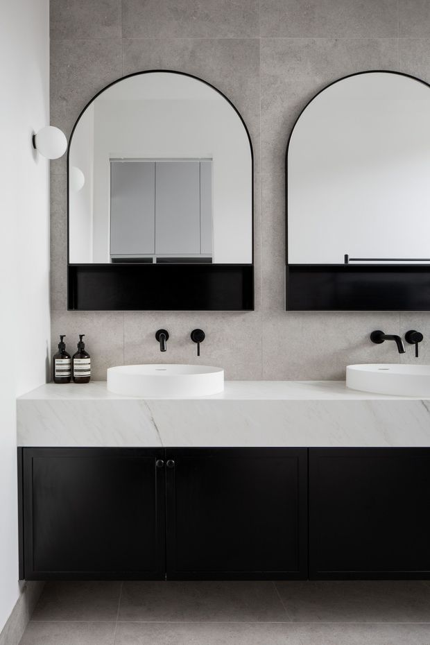 A contemporary double-vanity bathroom with two white countertop basins, black wall-mounted tapware, large arched mirrors in black frames, and a floating black vanity with a white veined stone benchtop. Large grey stone-look tiles cover the walls and floor, with minimal black-and-white styling and integrated round wall lights.