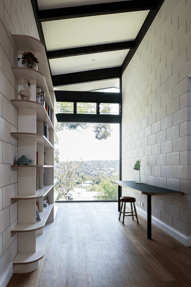 A narrow, light-filled study or home office with a long dark desk and stool beside a large window overlooking a leafy suburban landscape. One wall has white painted concrete blockwork, while the opposite wall features curved-edge built-in shelving and cabinetry. The ceiling has exposed black structural beams with translucent panels, creating a bright clerestory-like roof structure. The space has timber flooring and a minimalist, functional character.