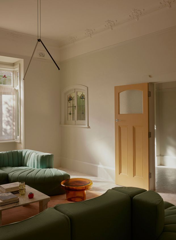 A warm, light-filled living room in a period home, featuring cream walls with ornate cornice detailing, a bay window, a small decorative stained-glass window, and an open painted timber door leading to an adjoining hallway or room. Deep green curved sofas, a translucent amber coffee table, and a low stone or tiled coffee table create a refined contemporary interior within the traditional architectural shell.