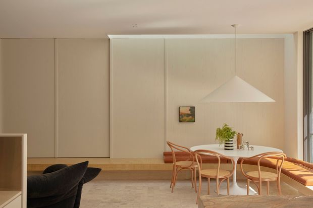 A warm, minimalist dining area with a round white dining table and curved timber chairs, set against pale timber wall panelling. A large white geometric pendant light hangs above the table, while a built-in timber bench runs along the wall beside full-height glazing. Dark sculptural lounge seating is partially visible in the foreground.