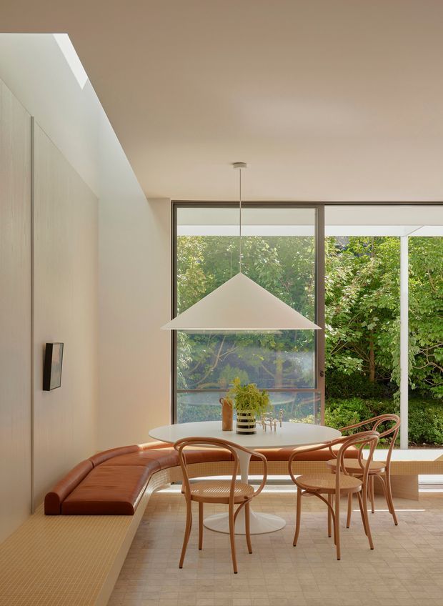 A bright, refined dining area with a built-in banquette along the left wall, a rounded white dining table, curved timber dining chairs, and a large geometric pendant light. Full-height glazing opens the room to dense greenery outside. The space features pale timber wall panelling, light tiled flooring, warm neutral finishes, and a contemporary mid-century-inspired furniture palette.