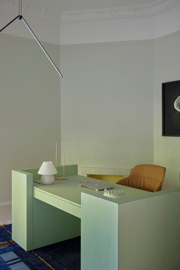 A refined home office in a heritage-style room, featuring a pale sage-green built-in desk with raised side panels and integrated cabinetry, a mustard leather office chair, a sculptural white table lamp, minimalist pendant lighting, and a dark patterned carpet. The room has pale green walls, ornate white ceiling cornices and decorative plaster mouldings, with framed artwork on the wall.