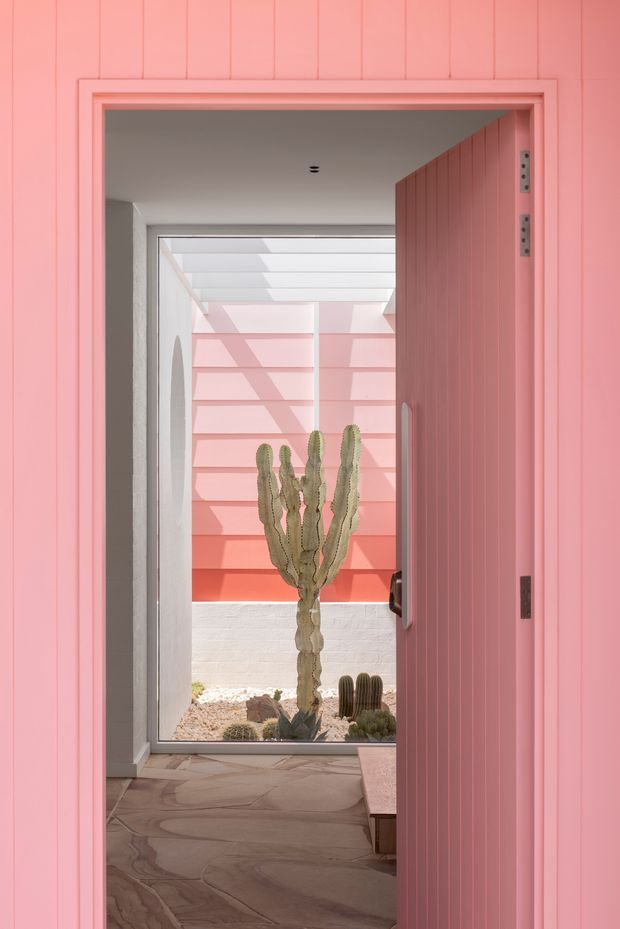 View through an open pink vertical-panel door into a bright, minimalist entry or passage. The space features a full-height glazed opening, a translucent roof or clerestory structure, pink exterior screening, and a small desert-style courtyard with sculptural cacti. The interior floor is finished in irregular natural stone, with strong pink and coral tones throughout.