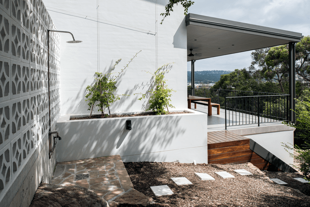 A modern outdoor courtyard and terrace beside a white-rendered building, featuring a decorative concrete block privacy wall, raised planter, exterior shower, gravel garden with stepping stones, timber decking, and a covered balcony overlooking a leafy hillside.