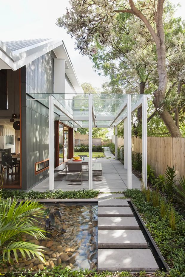 A bright contemporary residential courtyard viewed from an outdoor garden path. The scene features a modern extension with corrugated dark cladding, white structural framing, a translucent or glazed overhead courtyard canopy, and large glass openings. Concrete stepping slabs cross a shallow pebble-lined water feature, leading toward a paved outdoor dining area and landscaped rear garden. Mature trees, timber fencing, and lush planting create a leafy urban garden setting.