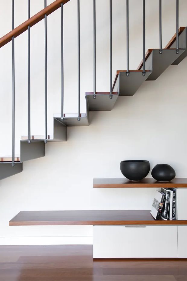 A minimalist interior featuring a floating staircase with grey metal treads, slim vertical dark metal balusters, and a warm timber handrail. Below the stairs is a streamlined built-in white cabinet with long timber shelves and dark decorative vessels, set against a white wall and warm timber flooring.