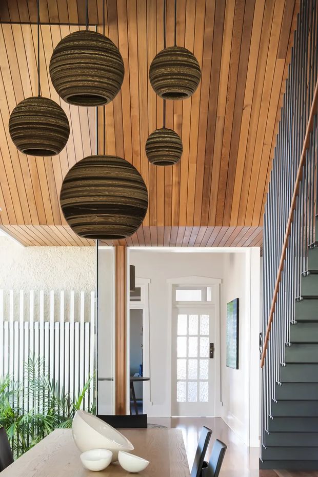 A bright contemporary entry and dining area featuring a warm timber-lined ceiling, five large woven spherical pendant lights, a white textured wall with vertical metal screening, and a dark open-riser staircase with timber handrail. A dining table with sculptural white bowls occupies the foreground, while a glazed front door and hallway are visible beyond.