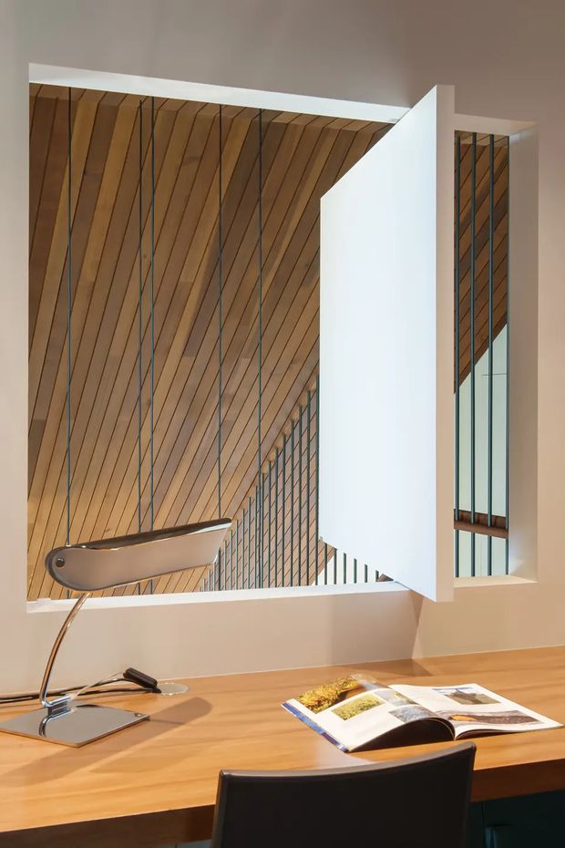 A refined home office or study with a timber desk, black desk chair, sculptural desk lamp and an open design book. A large internal architectural opening dominates the wall, revealing a dramatic timber-lined sloping ceiling, tall narrow glazing, and slender dark structural or window mullions. The space combines warm timber surfaces with crisp white geometric walls and a neutral plaster ceiling.