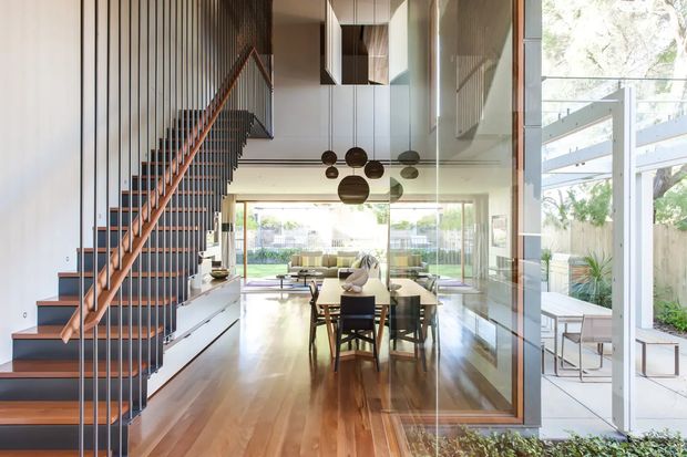 Bright contemporary open-plan interior showing a dining area connected to a living room and outdoor patio through extensive glazing. A sculptural timber-and-black metal staircase rises along a vertically slatted feature wall, while polished timber flooring, a long dining table, and clustered pendant lights define the central space. The room has a high ceiling and strong indoor-outdoor connection.