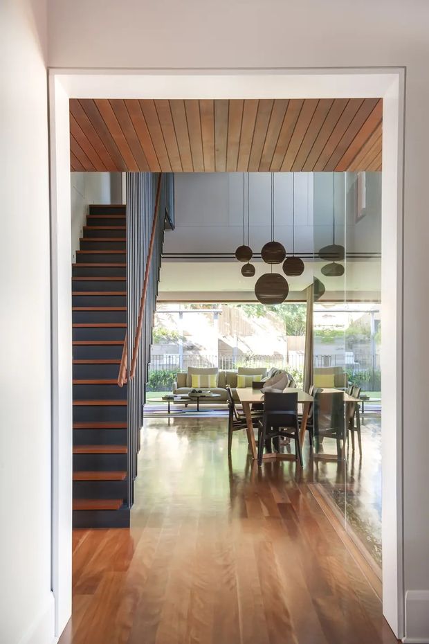 View through a wide interior doorway into an open-plan dining and living area. The space features polished warm timber flooring and a matching timber-lined ceiling, a dark open-riser staircase with timber treads and slim vertical balustrade, full-height glazing, a dining table with chairs, a low lounge seating area, and sculptural spherical pendant lights. The room is bright and refined with a strong indoor-outdoor connection.