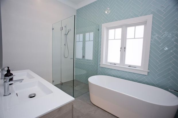 Bright contemporary bathroom featuring a freestanding white bathtub beneath a white-trimmed window, a glass-enclosed walk-in shower, and a double white vanity. The main feature wall is clad in pale blue-green herringbone tile, with light grey floor tiles and white walls and fixtures.