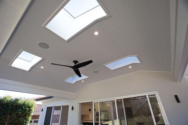 Covered outdoor entertaining area with a white-lined ceiling, multiple skylights, recessed lighting, and a central black ceiling fan. Large sliding glass doors open toward the house, with a small adjoining courtyard and garden visible beyond.