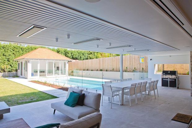 Bright covered outdoor entertaining area opening onto a swimming pool and landscaped lawn. The space features a large white louvred ceiling, concrete flooring, an outdoor dining setting, lounge seating, a built-in barbecue, timber screening, and a glass pool fence. A glazed pool pavilion is visible beyond.