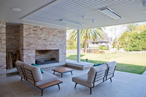 Covered outdoor living area with a large masonry fireplace, concrete floor, slatted ceiling, and comfortable lounge seating. The open-sided space overlooks a landscaped backyard and is designed for sheltered entertaining.
