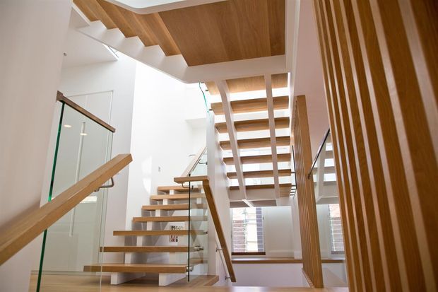 Bright contemporary stairway with open-riser timber stairs, glass balustrades, timber handrails, white walls, and a timber-lined ceiling. Vertical timber slat screening and large windows add architectural detail around the stair void.