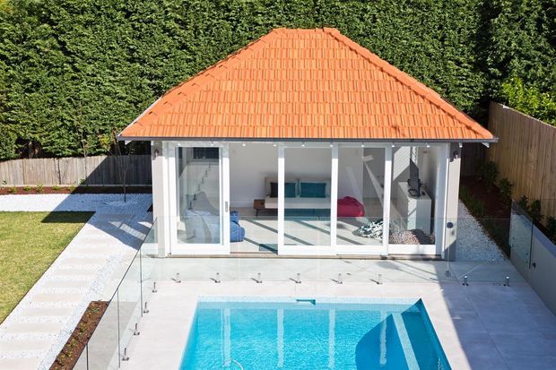 A bright contemporary backyard pool area viewed from above, featuring a rectangular swimming pool with glass safety balustrades and pale paving. Beyond the pool is a compact glazed pavilion or pool house with a terracotta tiled hipped roof, white walls, and full-height glass doors. Neatly landscaped garden beds, lawn, timber fencing, and tall hedging surround the space.