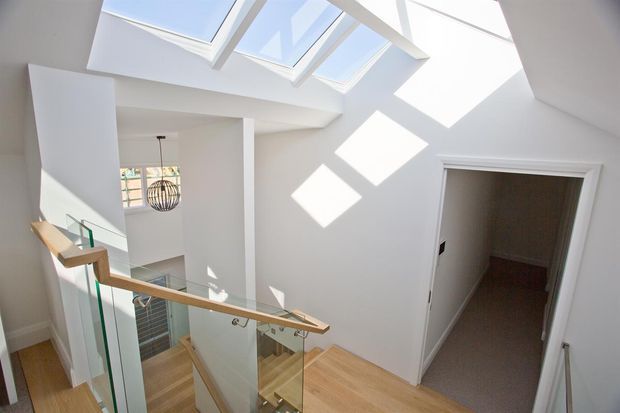 Bright contemporary stair landing with white walls, a timber handrail and timber flooring, and frameless glass balustrades. Multiple skylights flood the double-height stairwell with natural light, creating strong geometric shadows. A pendant light and glimpse of lower-level glazing are visible, along with an open doorway to a carpeted room.