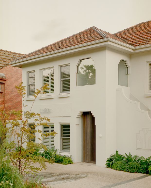 Exterior view of a renovated two-storey residence with a cream rendered facade, terracotta tiled pitched roof, detailed decorative window surrounds, timber vertical-slat entry door, plantation shutters, and an ornate curved exterior stair wall. Low garden planting and a pale textured driveway frame the frontage. The architecture combines traditional detailing with a clean contemporary renovation.