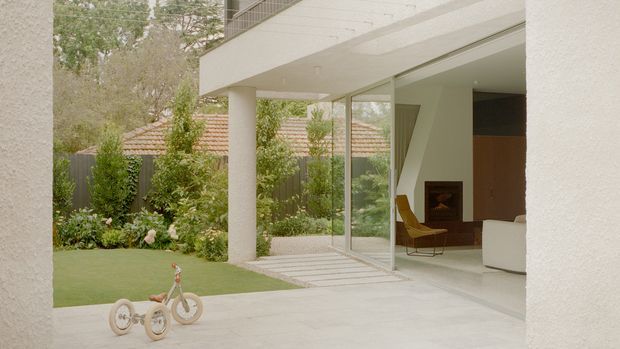 A modern residential courtyard and living area viewed from a covered outdoor space. Large frameless-looking glazed sliding doors open onto a pale paved terrace, revealing a minimalist interior with a sculptural white staircase, timber cabinetry, a fireplace, and a mid-century-style lounge chair. The exterior includes smooth white rendered walls, a broad flat overhang, lush planted garden beds, lawn, and a neighbouring terracotta tiled roof visible beyond the boundary.
