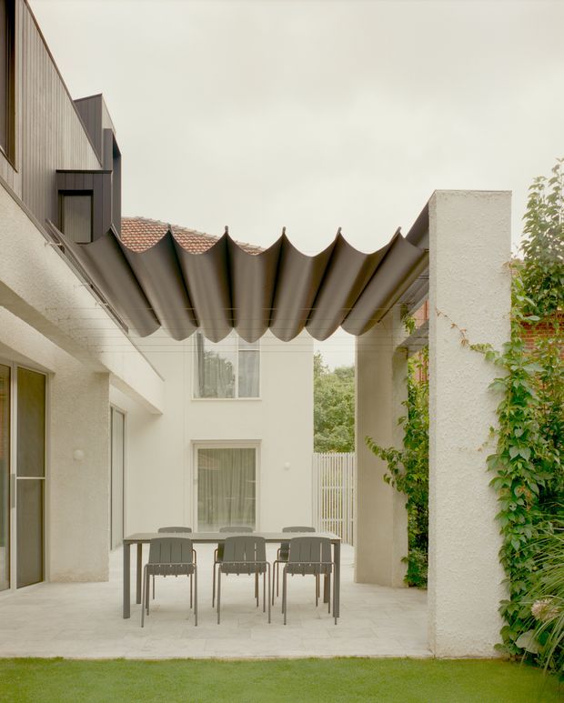 A sheltered residential courtyard or patio with a contemporary outdoor dining setting. A sculptural scalloped canopy spans between white textured walls, with a long outdoor table and six chairs on pale stone paving. Glazed doors and windows open onto the courtyard, while climbing greenery softens the perimeter and a small lawn occupies the foreground. The architecture combines clean geometric forms with textured rendered walls and a tiled roof visible beyond.