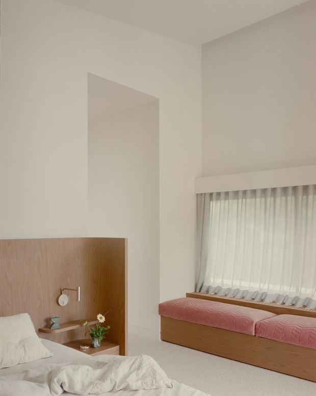 A softly lit minimalist bedroom with a low bed in the foreground and a built-in window seat along the right wall. The room features warm pale timber joinery, white walls and bedding, sheer full-height curtains, integrated bedside shelving, and a small wall-mounted reading light. Pink quilted cushions and a small vase of flowers add subtle colour.