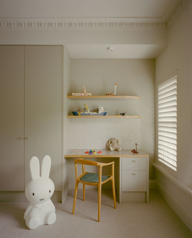 A warm, softly lit children's study nook or bedroom corner with built-in cabinetry, a compact desk, open display shelves, and a wooden chair. A large white rabbit toy sits on the carpeted floor, while white plantation shutters cover the window. The room features pale neutral finishes, simple decorative moulding, and playful children's accessories.
