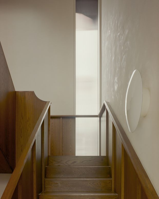 A narrow interior stairway viewed looking upward, with warm dark timber stair treads, timber balustrades and wall panelling. Soft daylight enters through a tall translucent vertical window at the landing, while a circular wall light or sculptural feature is mounted on the right wall. The composition is minimalist and refined, with strong vertical lines and warm natural materials.