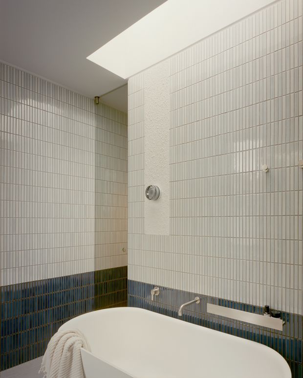 A compact, refined bathroom with a freestanding white bathtub in the foreground. The walls are clad in small vertically gridded white tiles with a contrasting band of deep blue tiles around the lower wall. A recessed skylight brings in soft natural light, while wall-mounted plumbing fixtures and a simple built-in ledge complete the space.