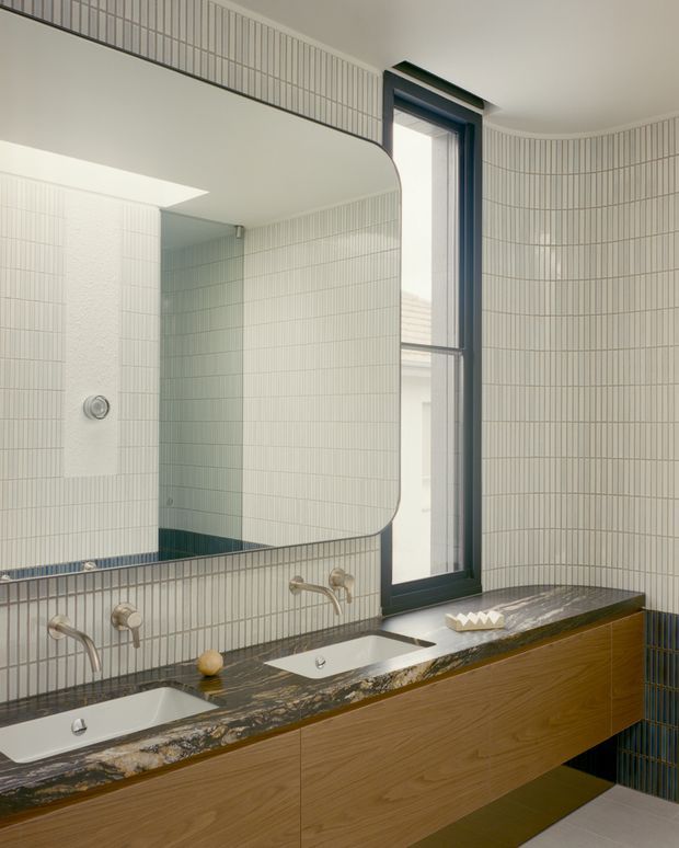 A refined bathroom featuring a long double vanity with integrated white basins, brushed metal wall-mounted taps, warm timber cabinetry, and a dark veined stone benchtop. Pale vertically arranged tiles cover the walls, with dark blue-green accent tiles at the lower wall and a tall dark-framed window. A large rounded-corner mirror spans the vanity, creating a bright, contemporary space.