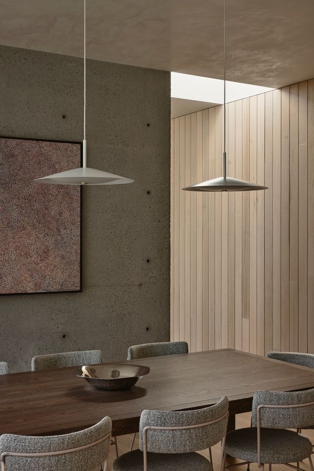 A refined dining room with a large dark timber dining table and upholstered dining chairs, framed by a raw textured concrete wall and vertical light timber wall panelling. Two slim sculptural pendant lights hang above the table, while a recessed ceiling opening introduces indirect daylight. A framed artwork and shallow decorative bowl add restrained accents. The palette is warm, earthy, and neutral with a contemporary architectural character.